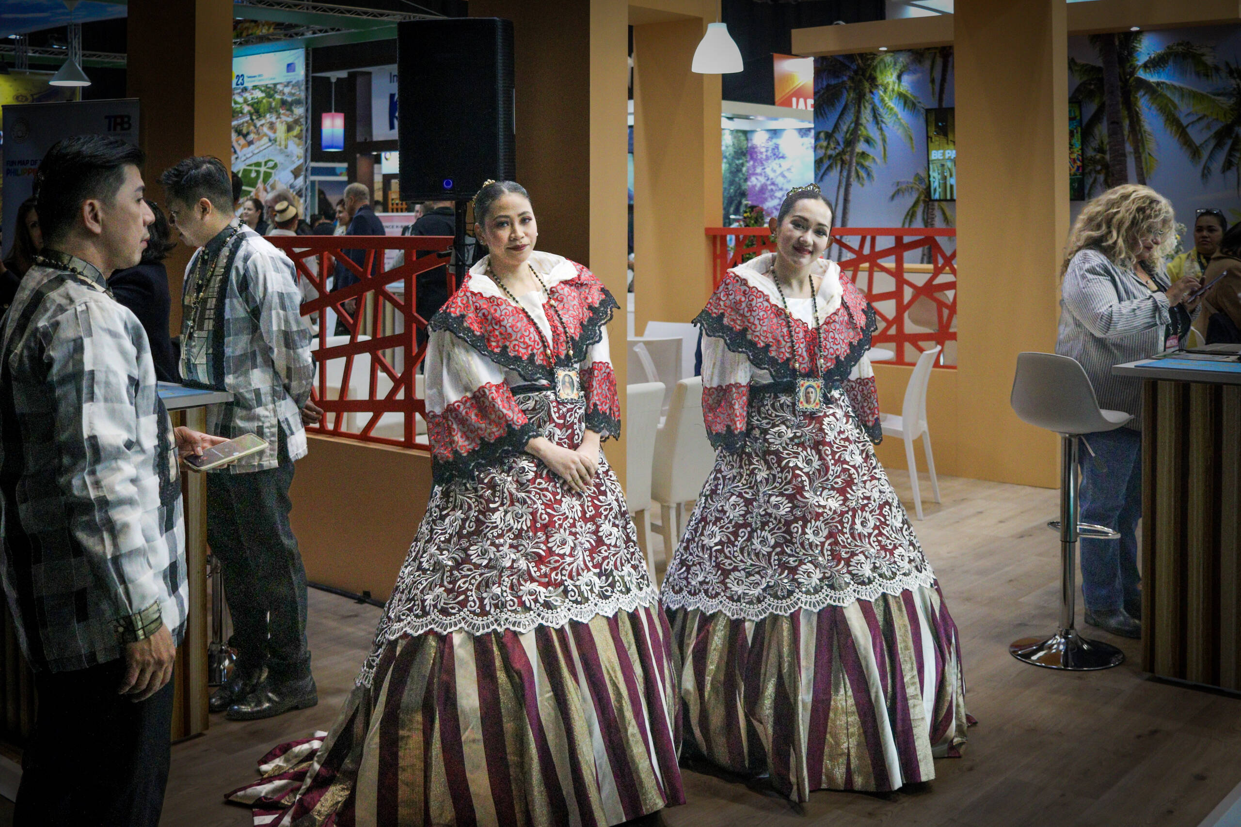 Two women in traditional Filipiniana gowns standing on a national tourist board stand at a travel trade fair