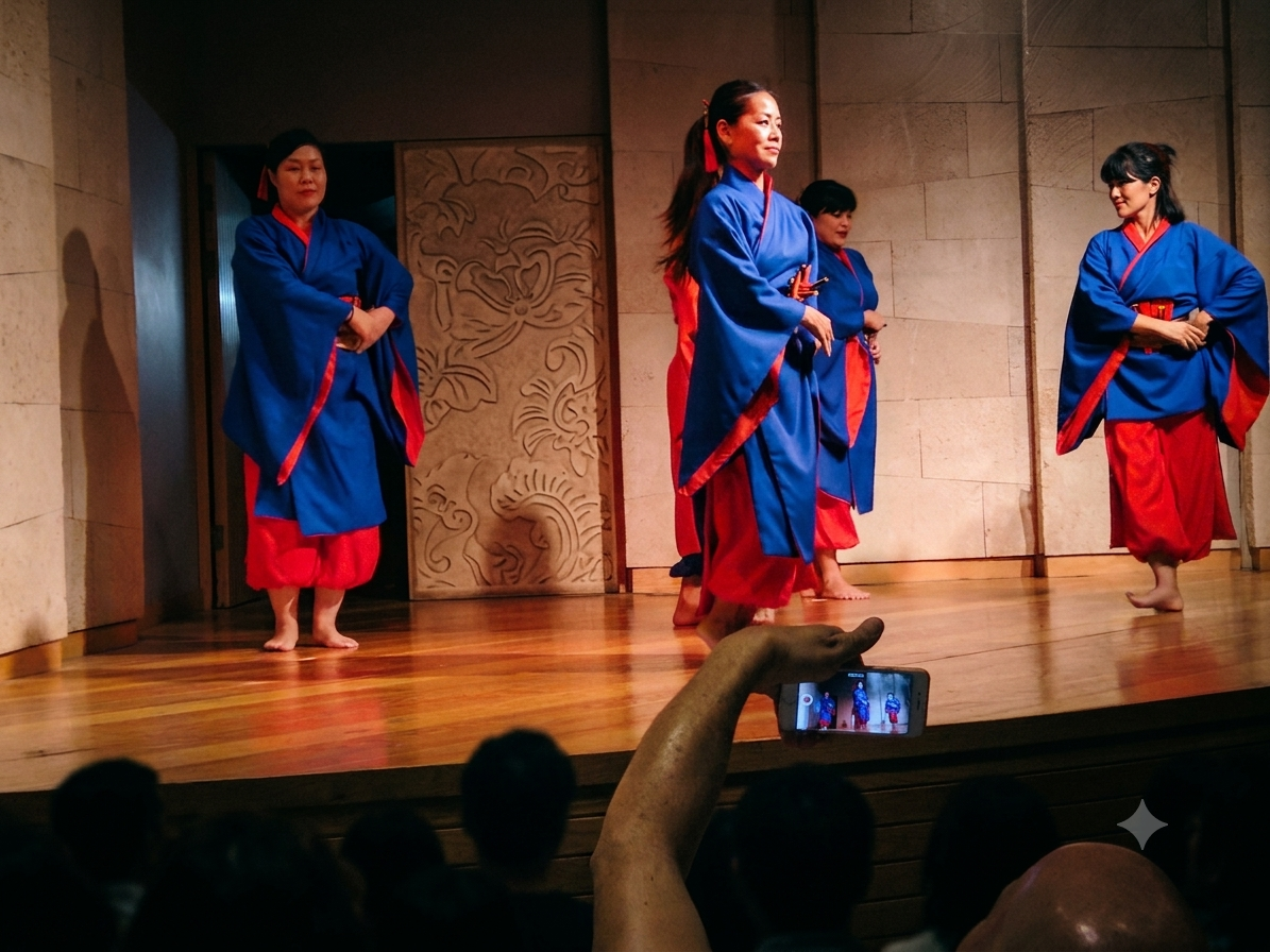 Four dancers in blue and red robes on a lit stage, dark auditorium and a raised phone in the foreground