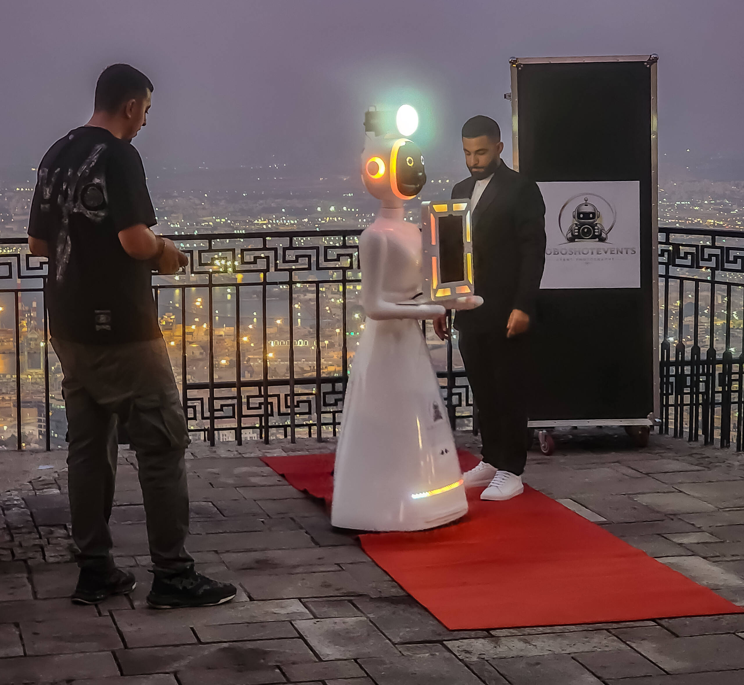 Event photo booth robot on a red carpet at a hilltop terrace during blue hour, with two operators and a city skyline behind