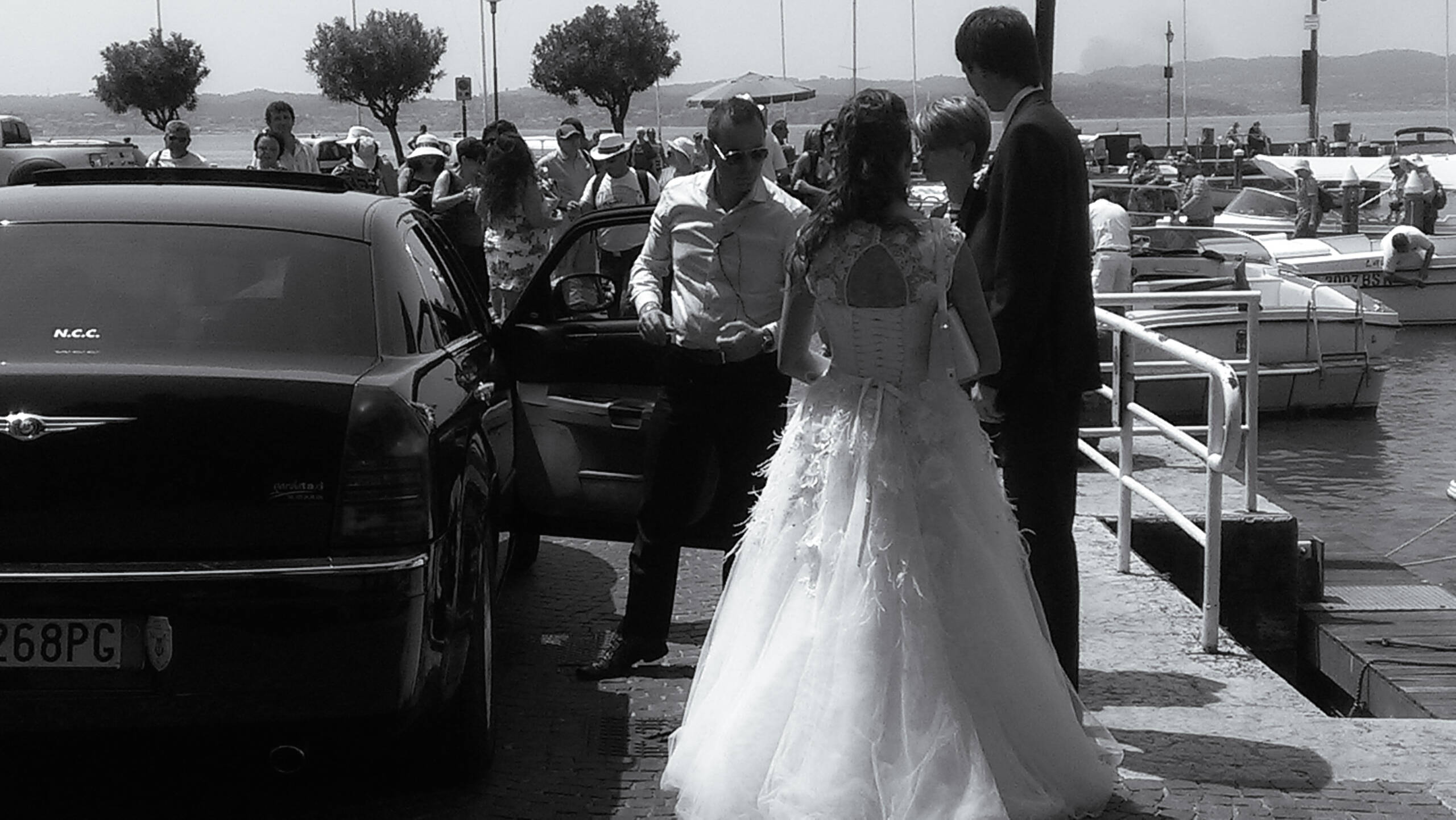 Black and white harbour frame: a bride in a full lace gown crossing cobbles toward a black chauffeur-driven saloon, guests and moored boats behind, hard midday sun