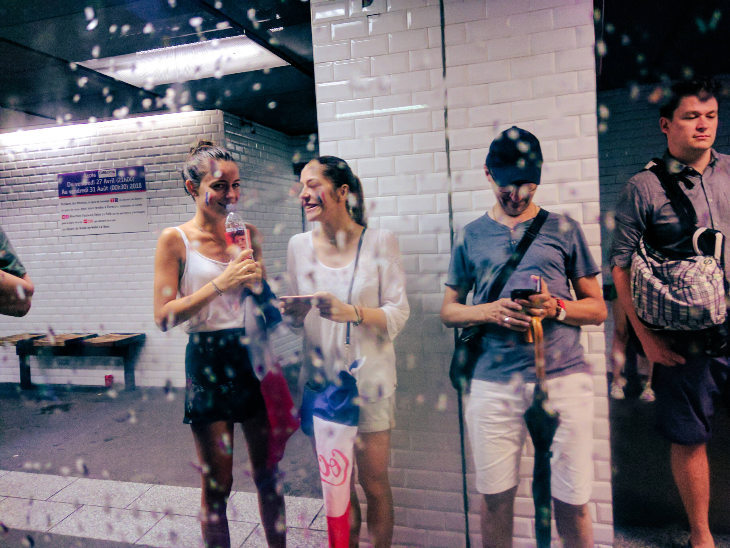 Fans with tricolour face paint on a Lyon metro platform, shot from inside an arriving train through a rain-covered window