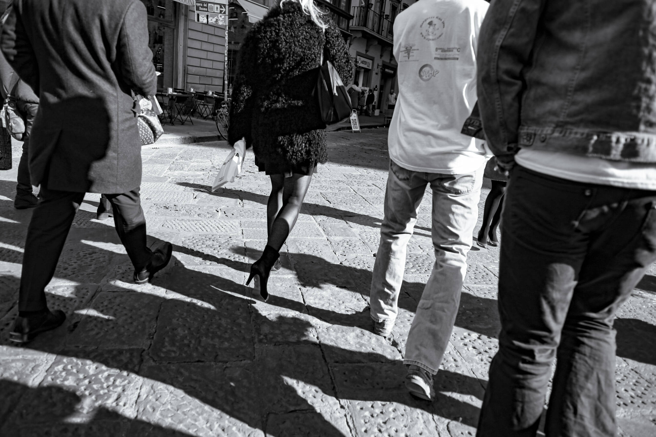 Pedestrians crossing a street in Florence, shot from behind at waist height, hard shadows across the paving