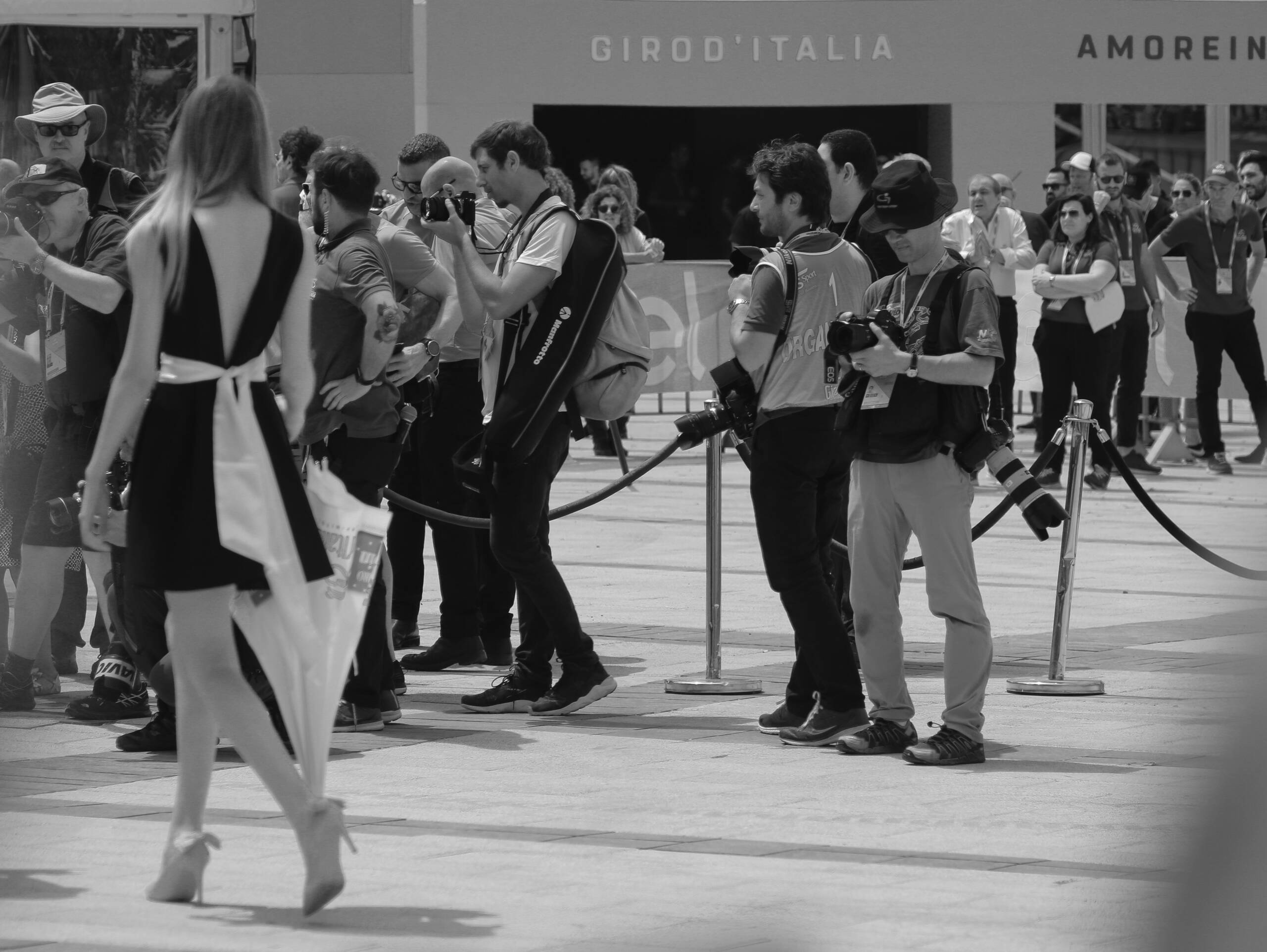 Press photographers in accreditation tabards at the Giro d&rsquo;Italia, ignoring a woman walking past the rope line