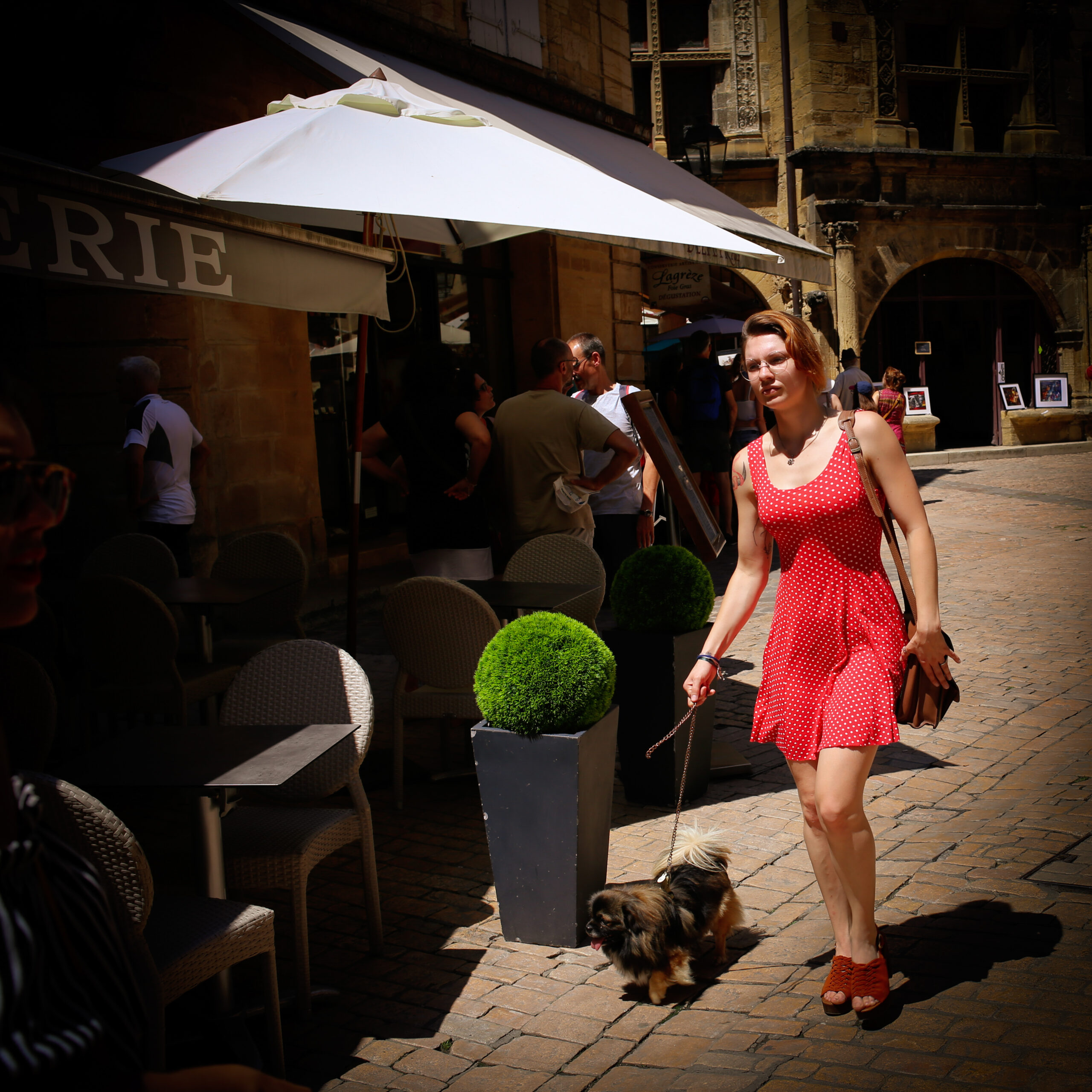 Woman in a red polka-dot dress walking a Pekingese past a café terrace in Sarlat